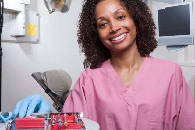 A dental assistant is wearing a pink shirt and is in the dental room